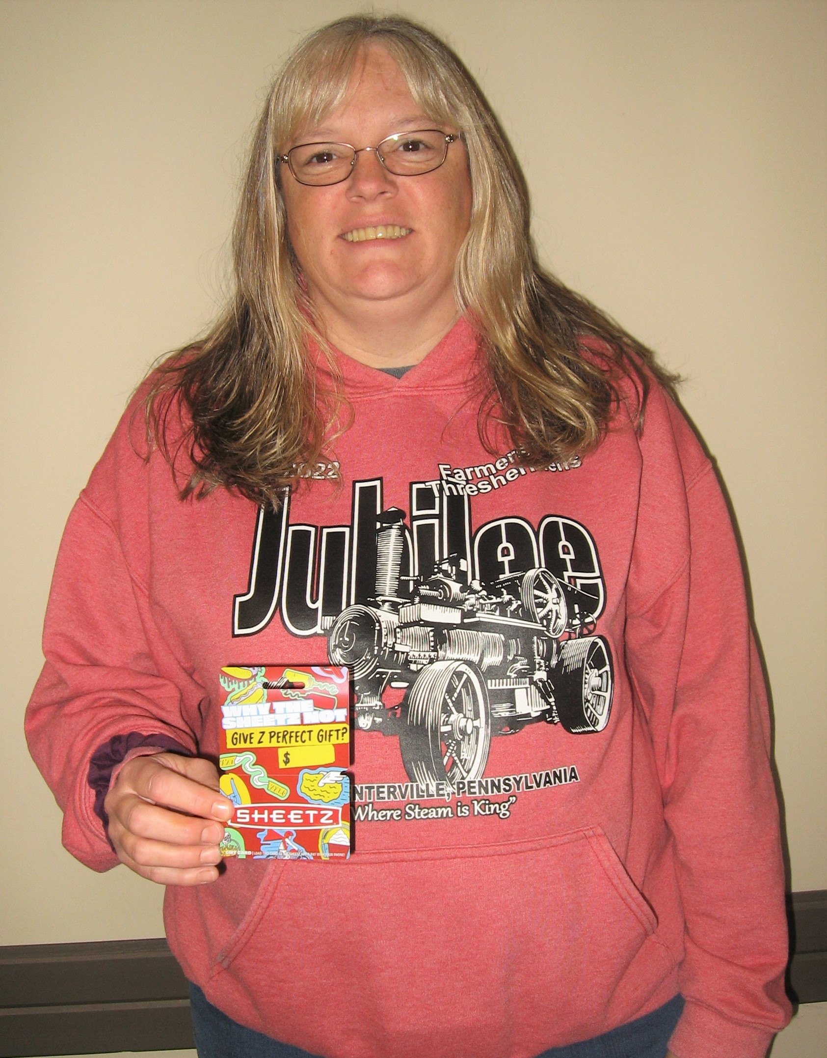 An older blonde woman with glasses and a pink shirt, holding a gift card