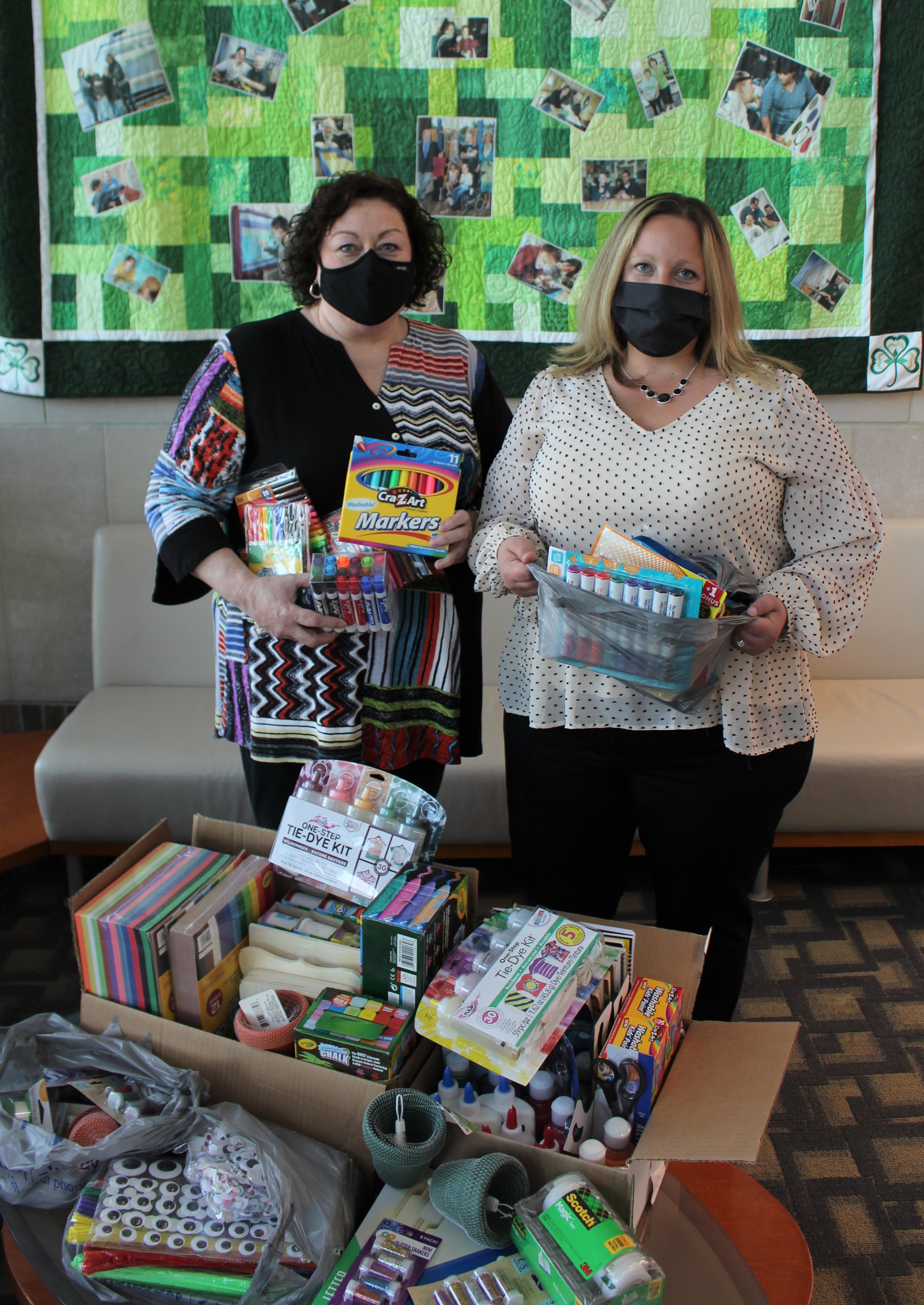 Two women wearing face masks, standing in front of a donated box of markers, paint, and other craft supplies. They're both holding markers